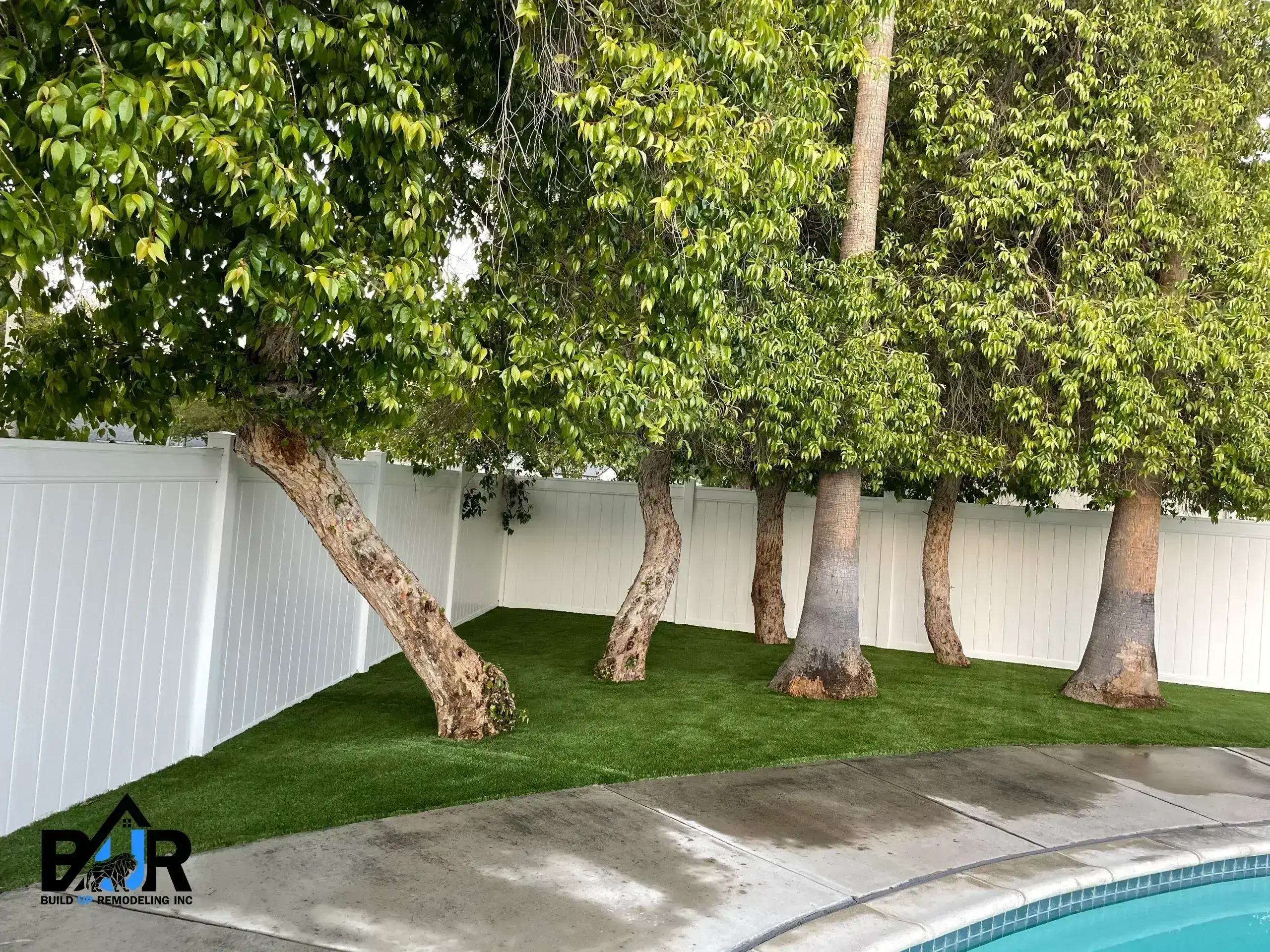 Curved tree trunks near a pool with artificial grass and white fencing.